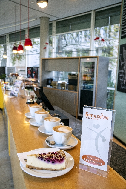 Theke des Cafés im Museum für Kommunikation Frankfurt mit einem Stück Käse-Beeren-Kuchen im Vordergrund und vier Tassen Cappucino aufgereiht dahinter