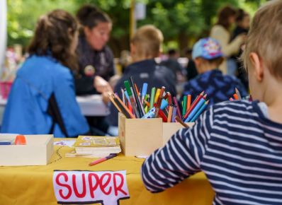 Ein Kind von hinten fotografiert, sitzt an einem Tisch mit gelber Tischdecke im Freien und malt mit Buntstiften, im Hintergrund weitere Kinder und Personen