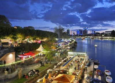 Buden und Bühnen mit erleuchtetem Boot am Main bei Nacht und Besucher*innen des Museumsuferfests