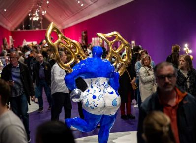 Viele Menschen in einem Ausstellungsraum, in dem in der Mitte eine blau-weiße abstrakte Skulptur einer rundlichen Frau mit goldenen Flügeln steht