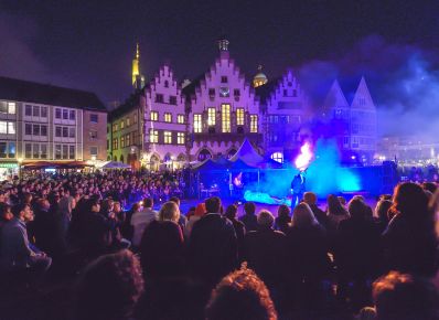 Auf dem Platz vor dem Frankfurter Rathaus bei Nacht stehen Zelte und ein Feuerschlucker in der Mitte, darum sitzen und stehen viele Menschen und schauen zu